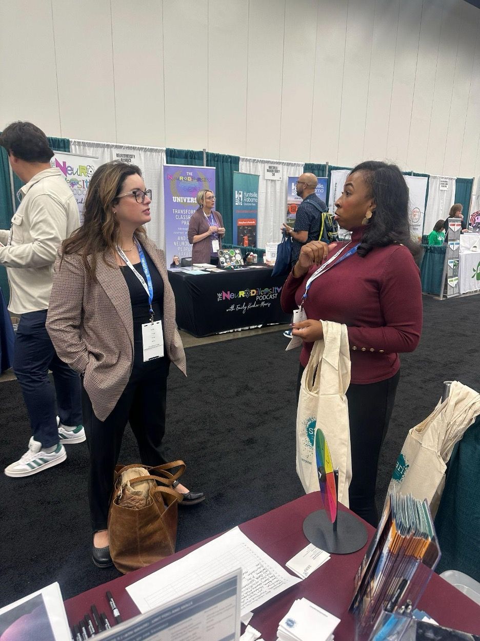 Two women converse at a trade show booth, holding a tote bag. Others browse exhibits.
