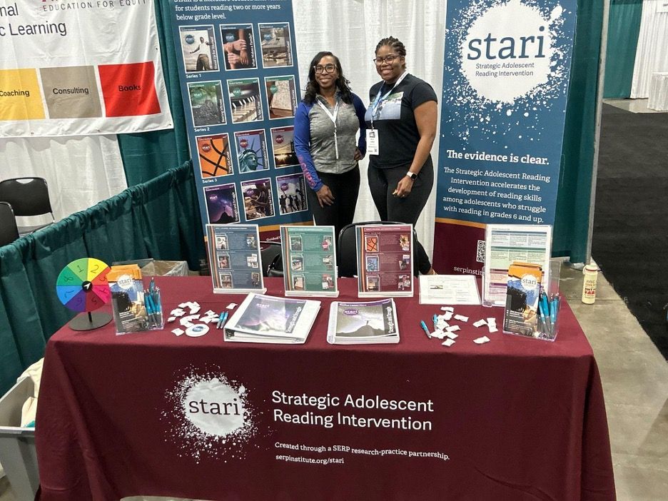 Two people stand at a trade show booth with brochures and a banner that reads