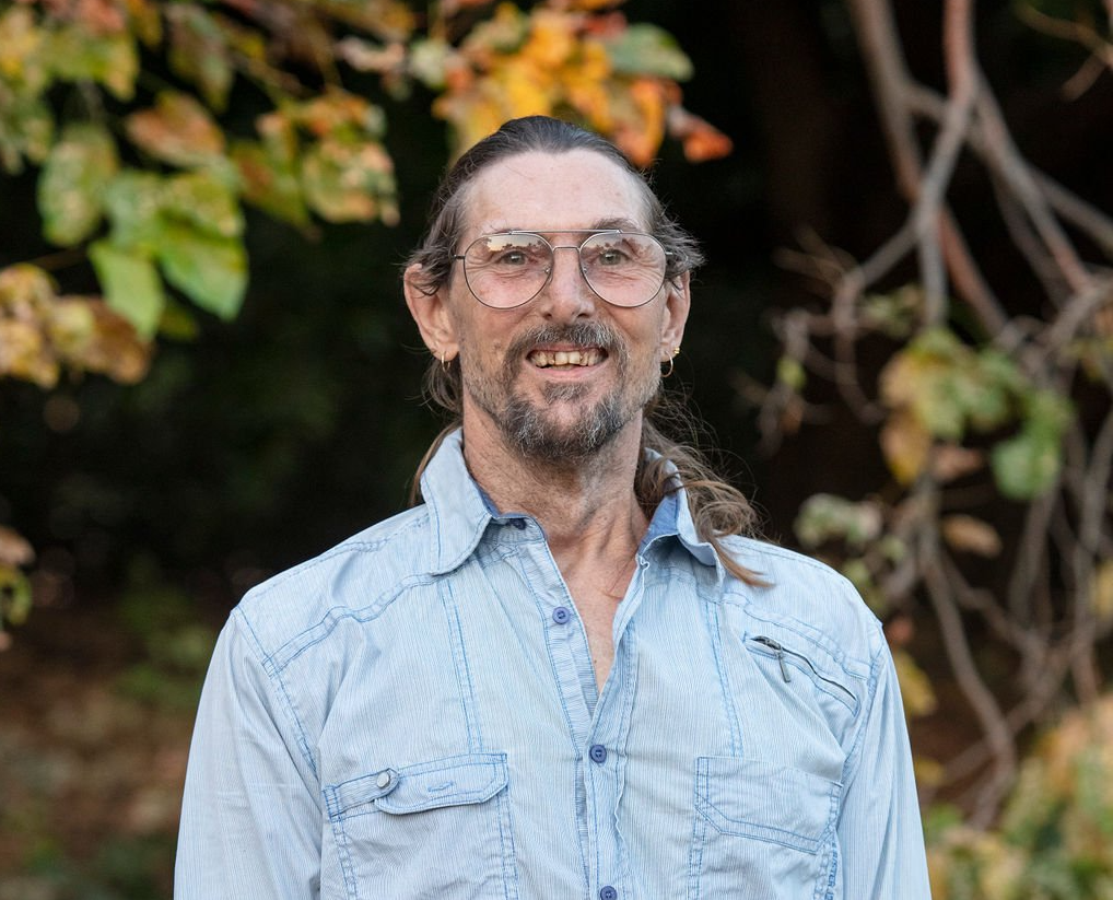 A man wearing glasses and a blue shirt is standing in front of a tree.