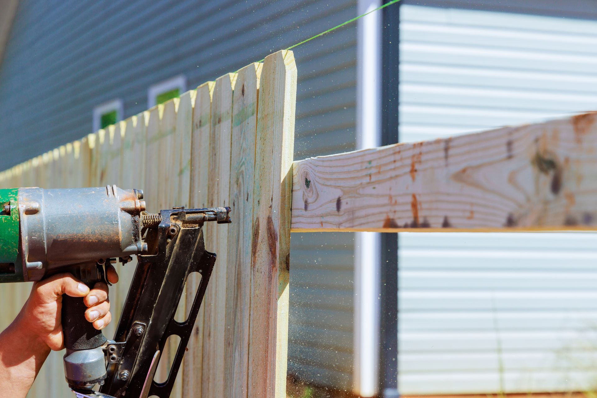Person using a nail gun to attach a wooden fence board to a horizontal beam outdoors.