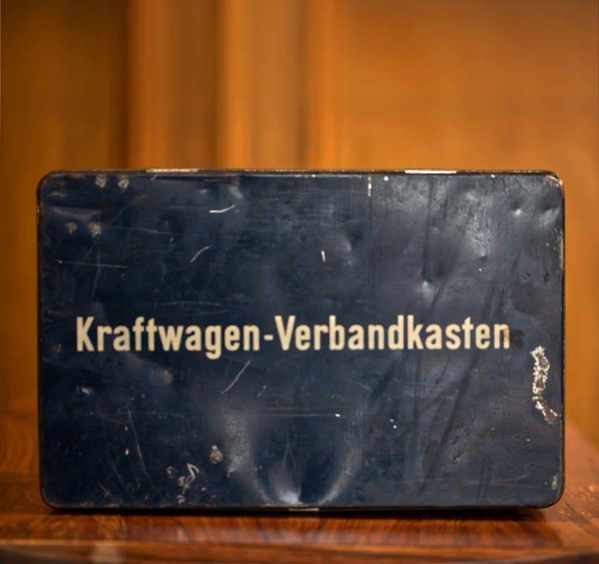 Ein alter, abgenutzter, schwarzer Metall-Erste-Hilfe-Kasten mit der Aufschrift „Kraftwagen-Verbandkasten“ in weißer Schrift, der auf einer Holzoberfläche steht.