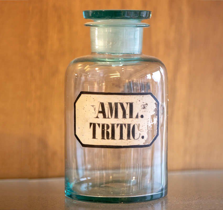 Eine leere, antike Apothekerflasche aus Klarglas mit Stöpsel, beschriftet mit „AMYL. TRITIC.“ in schwarzer Schrift.