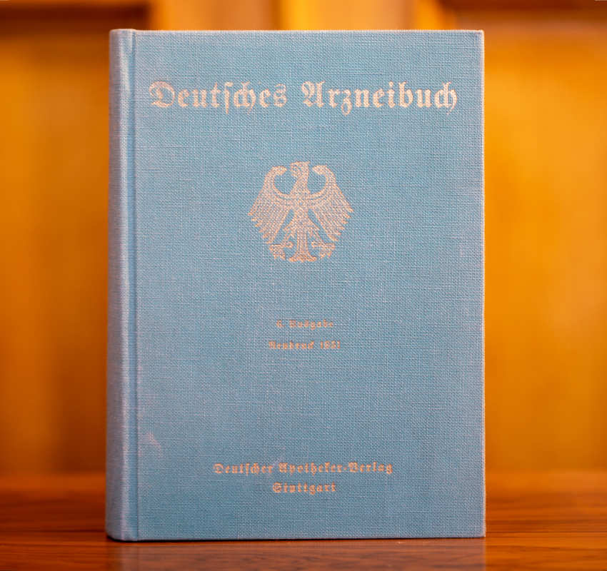 Ein blaues Buch mit dem Titel „Deutsches Arzneibuch“, das einen aufrecht auf einer Holzoberfläche stehenden deutschen Adler zeigt.