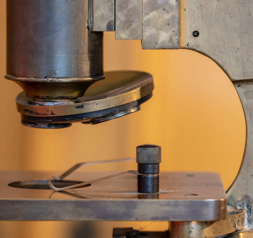 Nahaufnahme eines antiken Messingmikroskops, die die über einem Metalltisch mit Federklemme positionierte Objektivlinse zeigt.