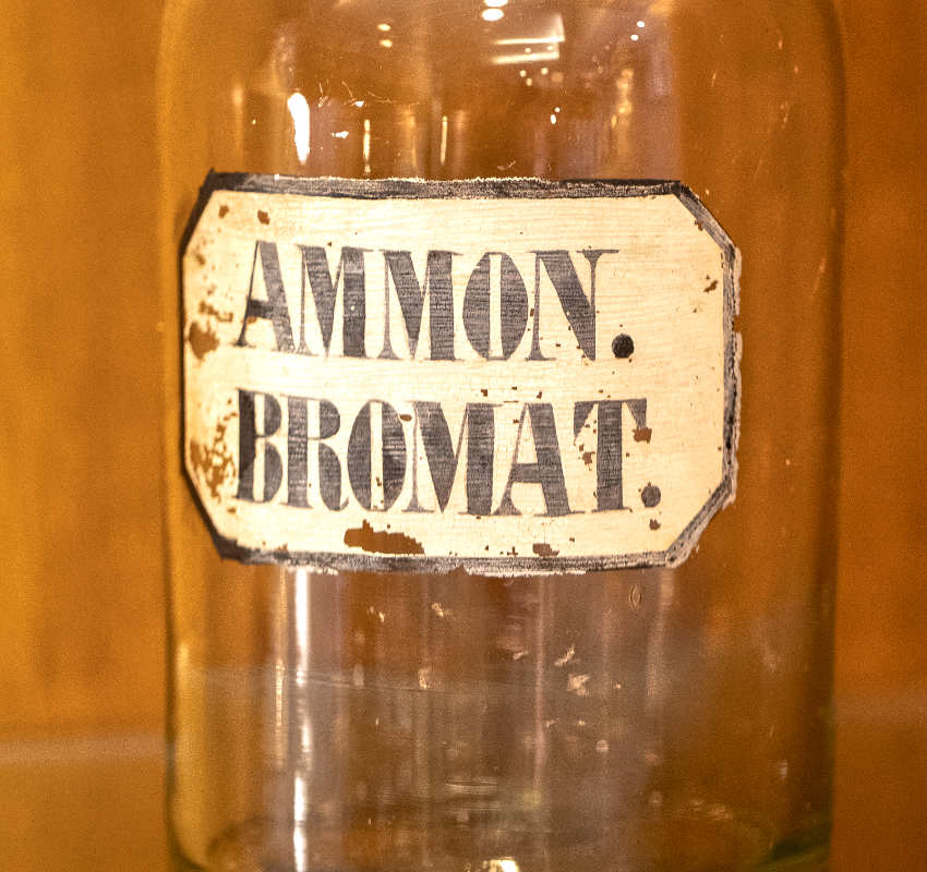 Eine durchsichtige Apothekerflasche aus Glas mit einem Papieretikett, auf dem „AMMON. BROMAT.“ steht vor einem schlichten Holzgrund.
