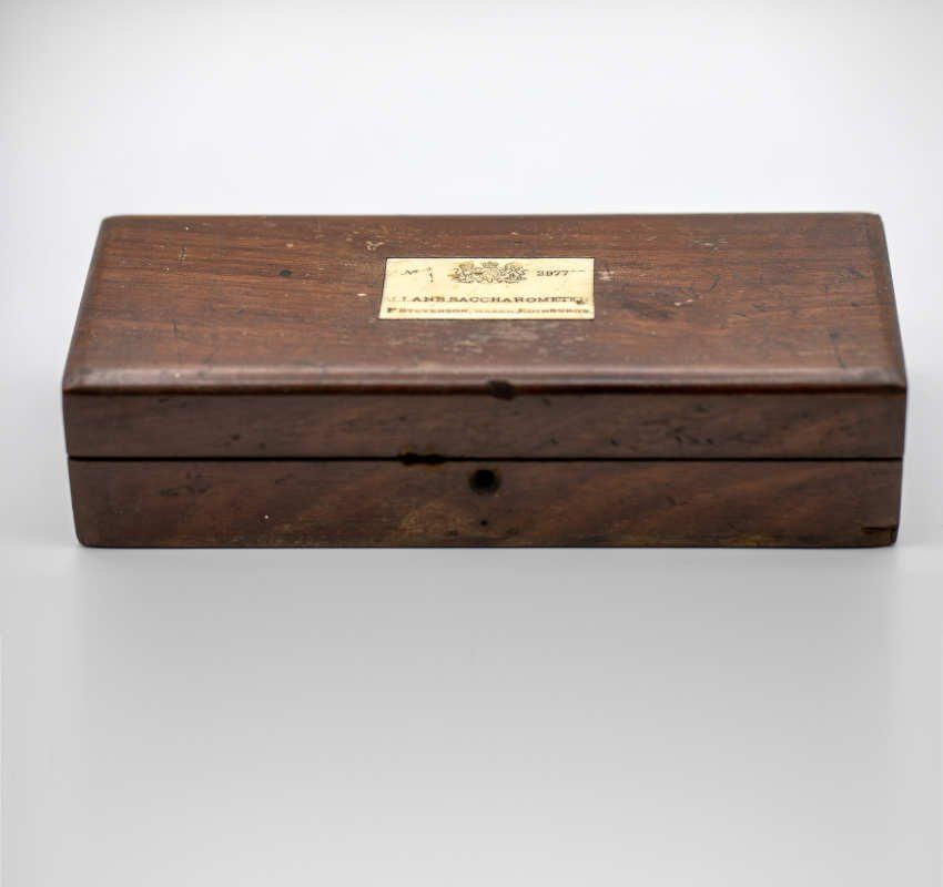 Eine dunkle, rechteckige Holzbox mit einem mittig platzierten weißen Etikett, die auf einer schlichten, hellen Oberfläche steht.