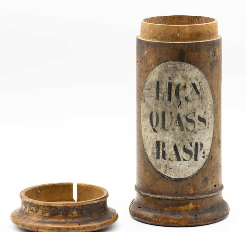 Ein antikes Apothekergefäß aus Holz mit separatem Deckel, beschriftet mit „LIGN QUASS RASP“ auf einem weißen ovalen Papieretikett.