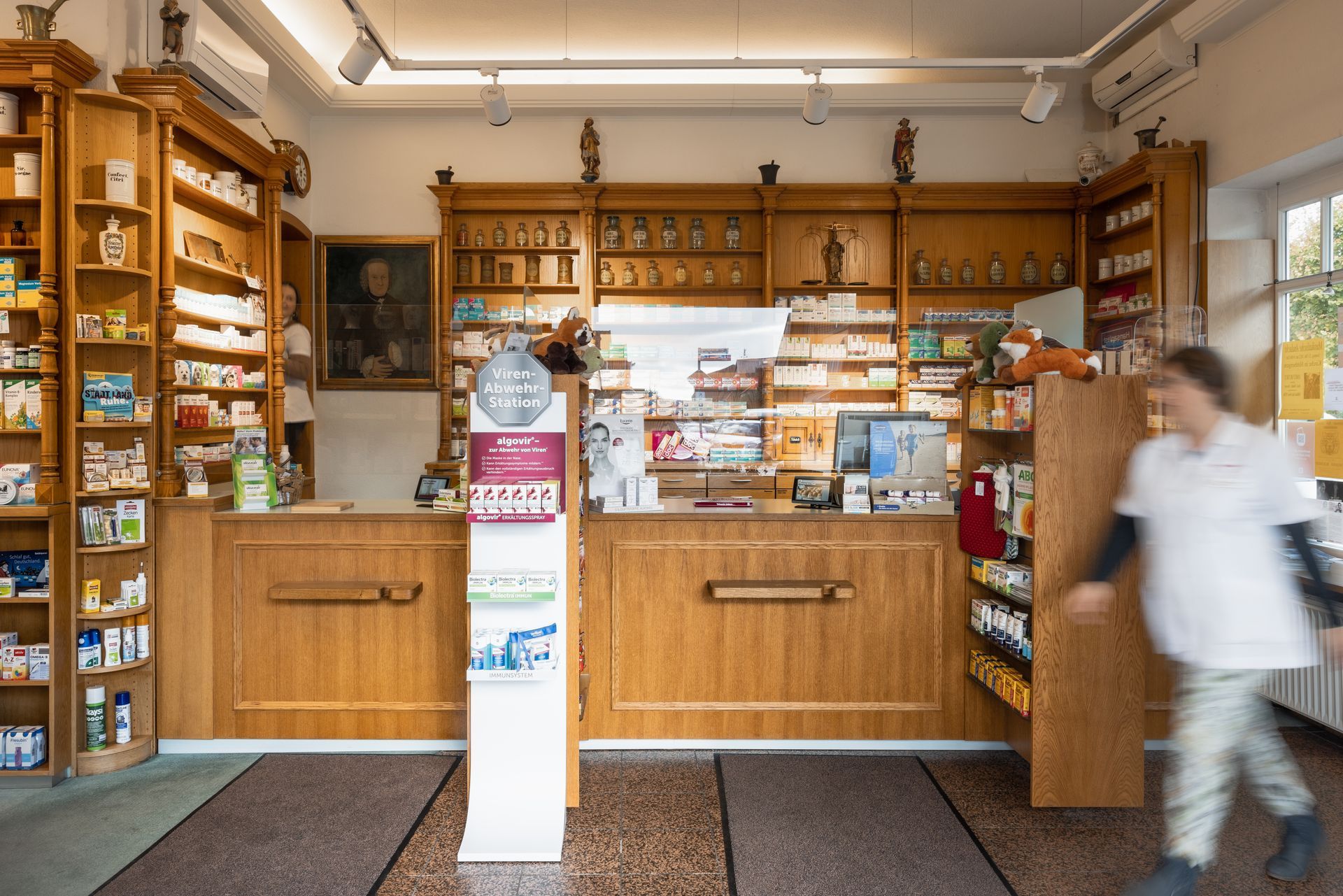 Eine Apotheke mit Holzregalen, auf denen Produkte ausgestellt sind, einem Bedienschalter und einer verschwommenen Person, die im Vordergrund vorbeigeht.