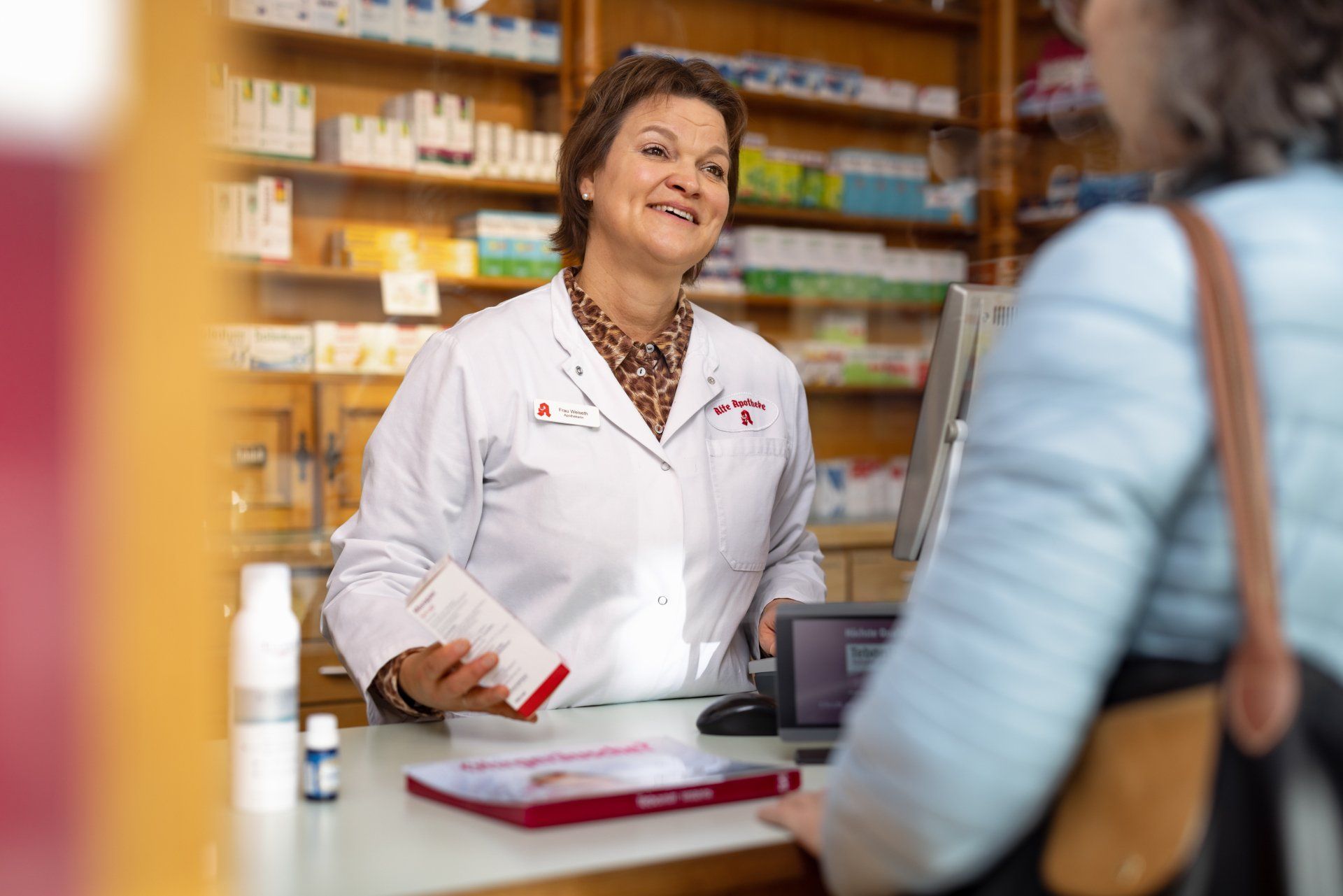 Ein Apotheker im weißen Kittel lächelt, während er einem Kunden am Apothekentresen eine Produktverpackung hinhält.