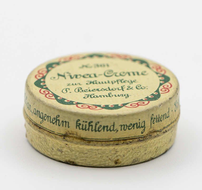 Eine runde, cremefarbene Nivea Creme Dose im Vintage-Stil mit dekorativen grünen und roten Rändern auf schlichtem weißen Hintergrund.