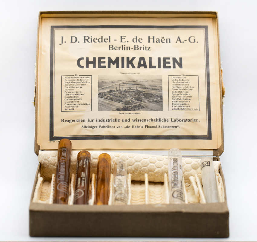Eine geöffnete Vintage-Chemie-Set-Box mit Glasreagenzgläsern und einem historischen Etikett von JD Riedel – E. de Haën A.-G.