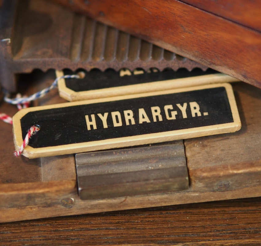 Ein Etikett mit der Aufschrift „HYDRARGYR.“ in hellen Blockbuchstaben auf schwarzem Hintergrund, das in einer alten Holzkiste liegt.