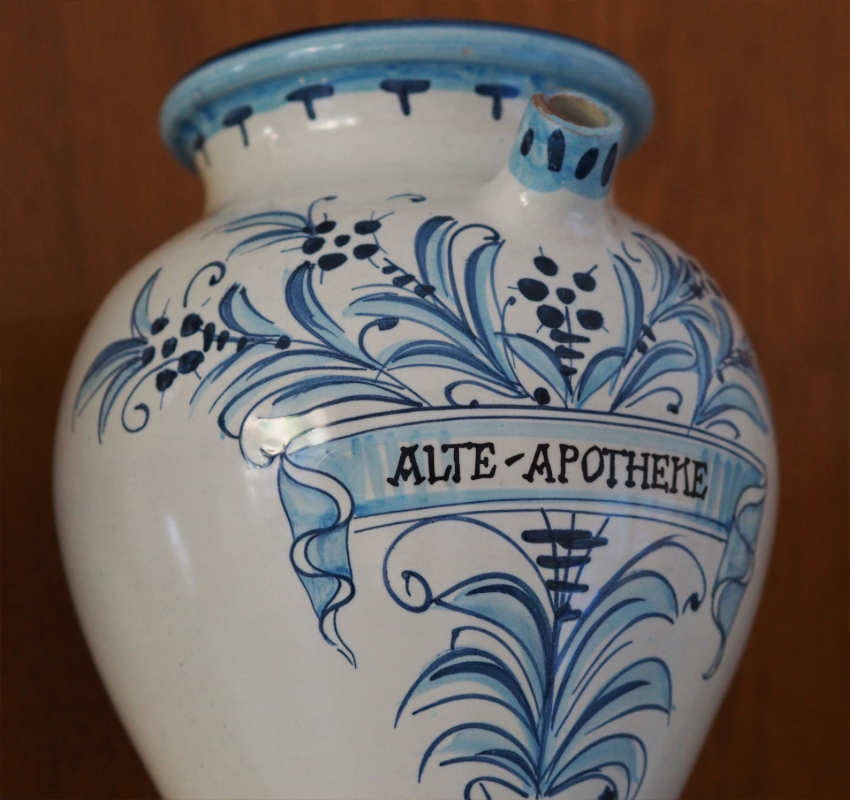 Ein weißes Apothekergefäß aus Keramik mit blauen Blumenmustern und dem Schriftzug „ALTE-APOTHEKE“ auf einem zentralen Banner.