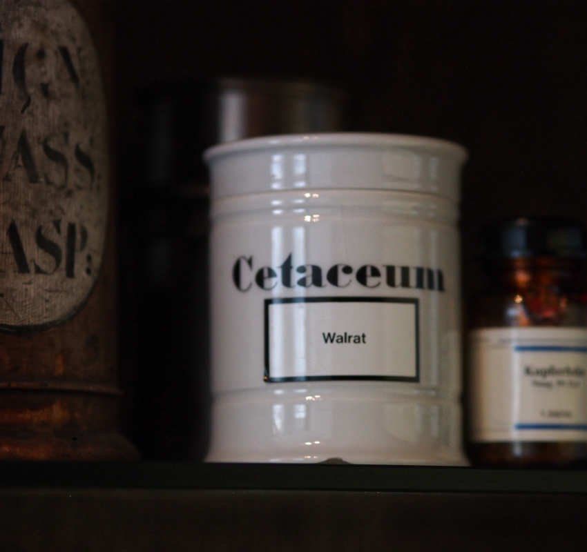 Ein weißes Apothekerglas mit der Aufschrift „Cetaceum“ und „Walrat“ steht auf einem Regal zwischen zwei anderen Behältern.