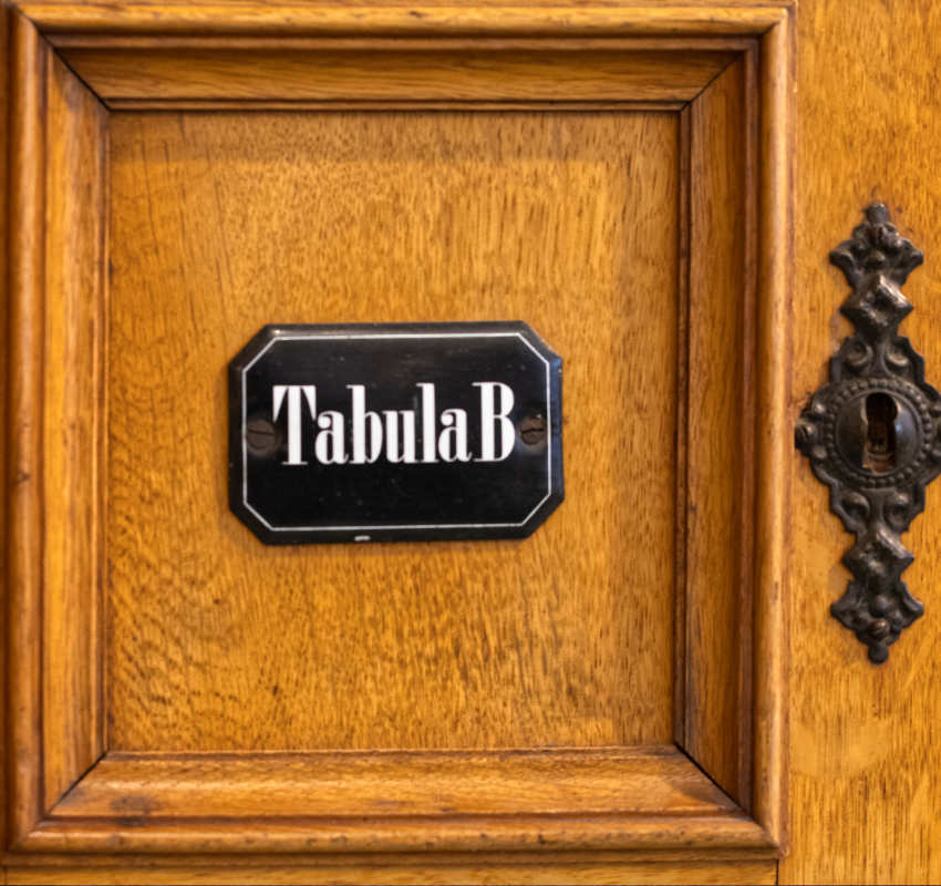 Nahaufnahme eines hölzernen Türblatts mit einer schwarzen rechteckigen Plakette mit der weißen Aufschrift „Tabula B“ und einem eisernen Schlüsselloch.
