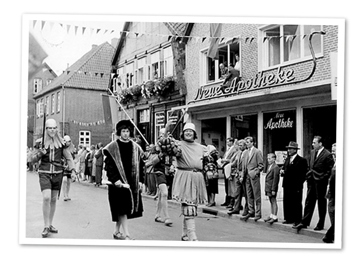 Während einer Parade marschieren Menschen in historischen Kostümen eine Straße entlang an einem Gebäude mit der Aufschrift „Neue Apotheke“ vorbei.