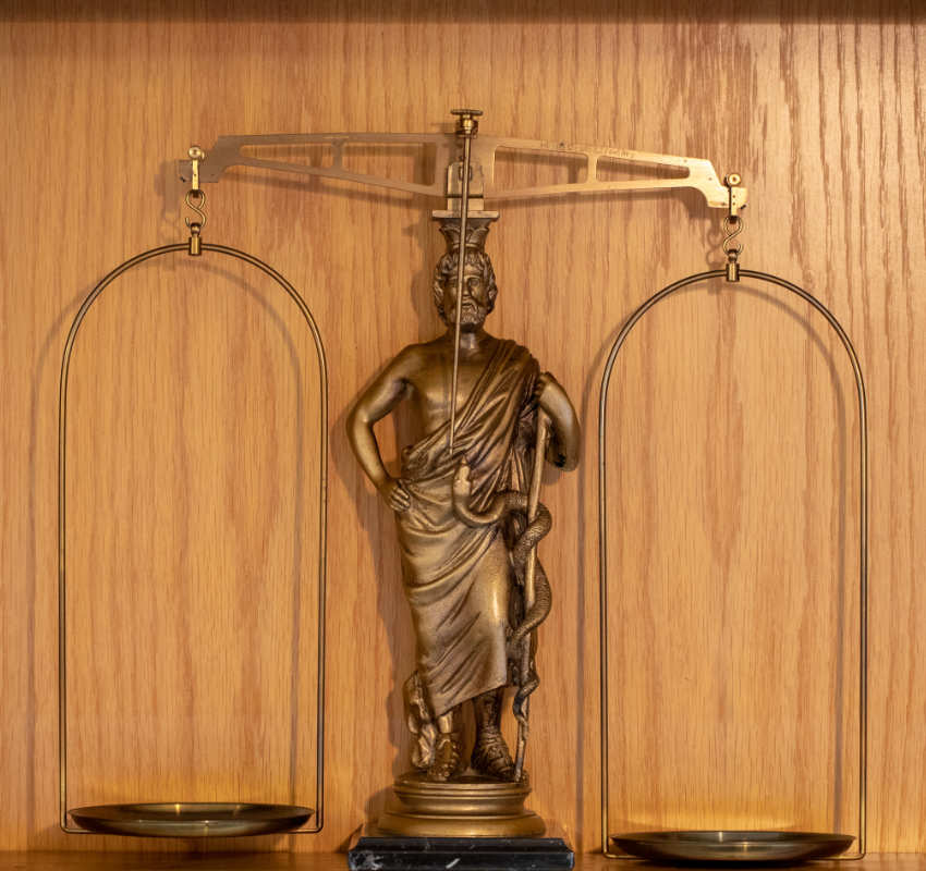 Eine bronzefarbene Statue einer Figur, die einen Stab mit einer zusammengerollten Schlange hält, zentriert unter einer ausbalancierten Waage vor einem Holzvorhang.