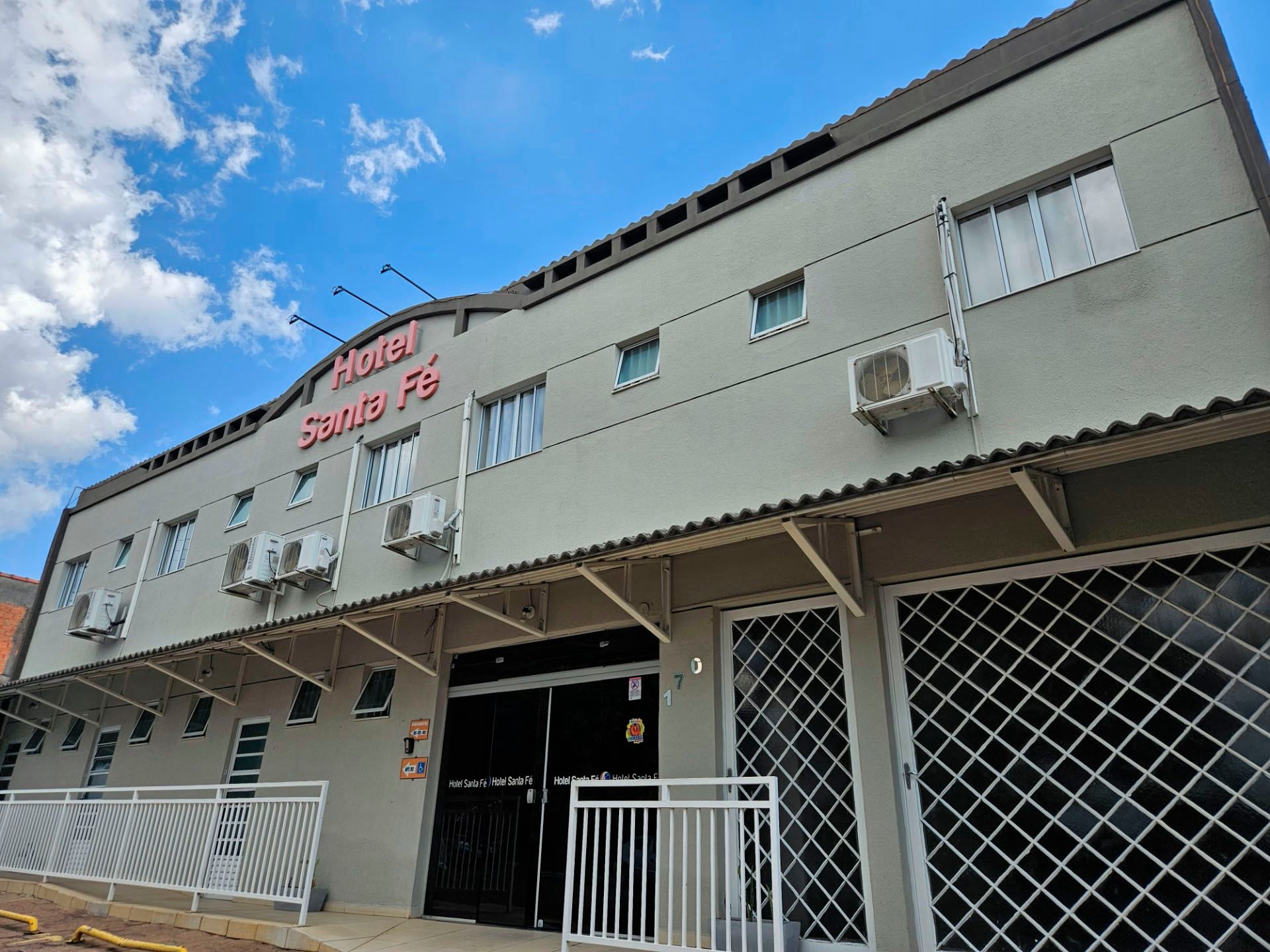 Exterior de um hotel bege de dois andares, com uma placa vermelha, toldo metálico e grades brancas sob um céu azul