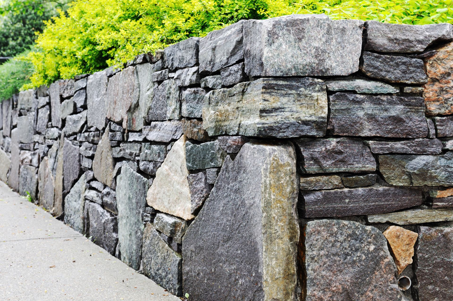 A stone wall along a sidewalk with trees in the background.