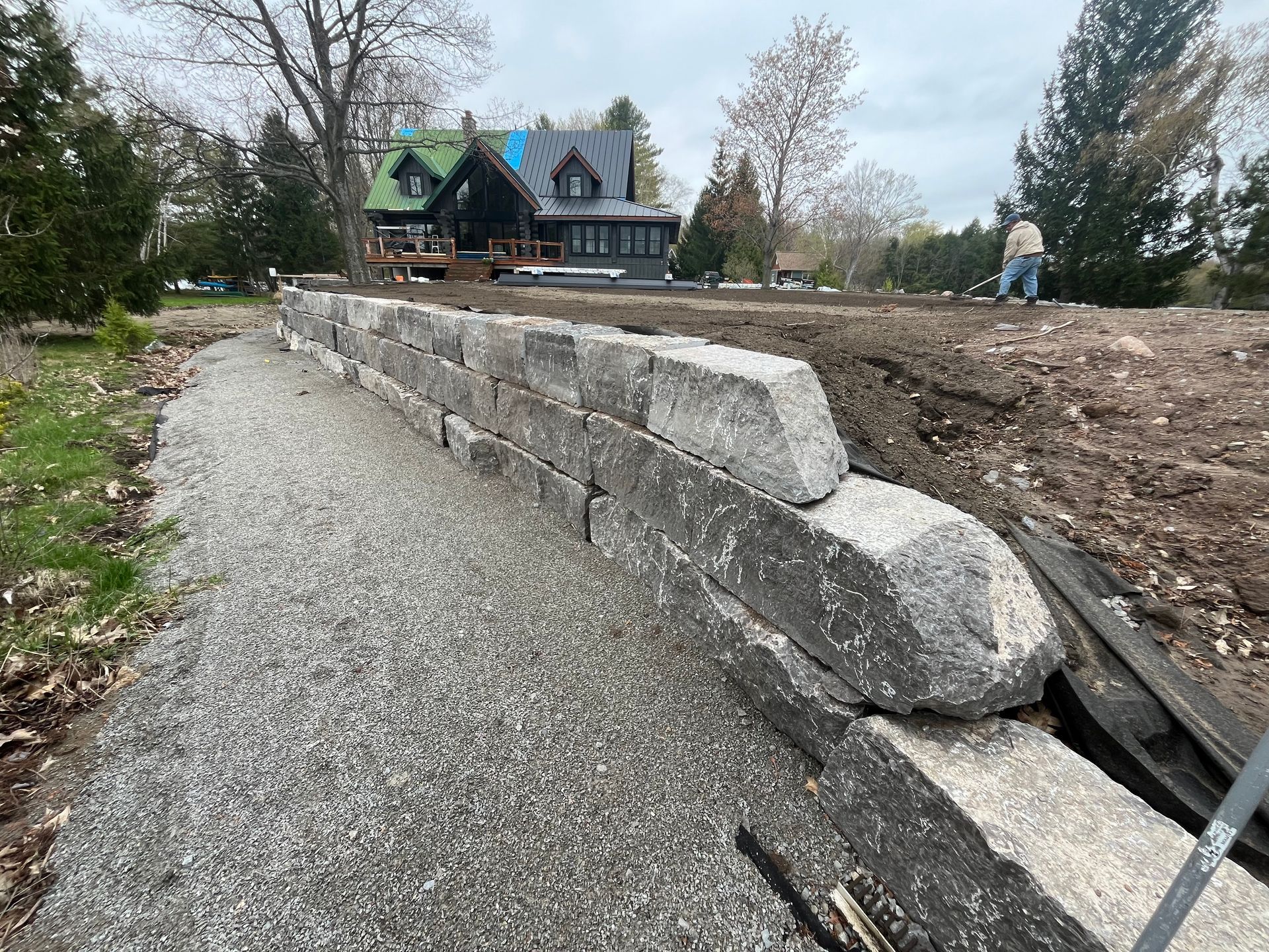 A retaining wall made of large stone blocks under construction; gravel in foreground; house in background.