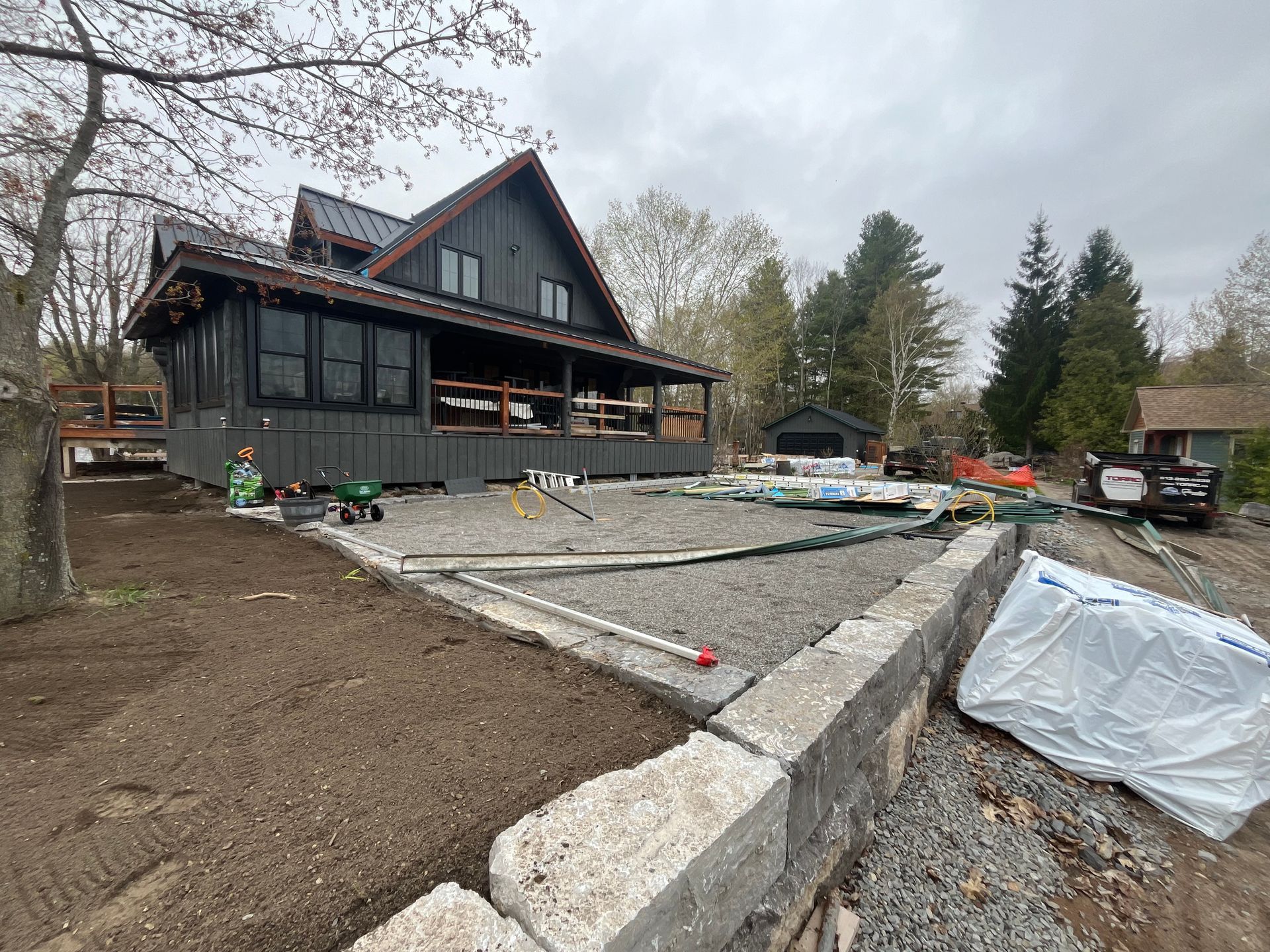 Dark, modern house under construction near water, with gravel and retaining wall in foreground. Cloudy day.