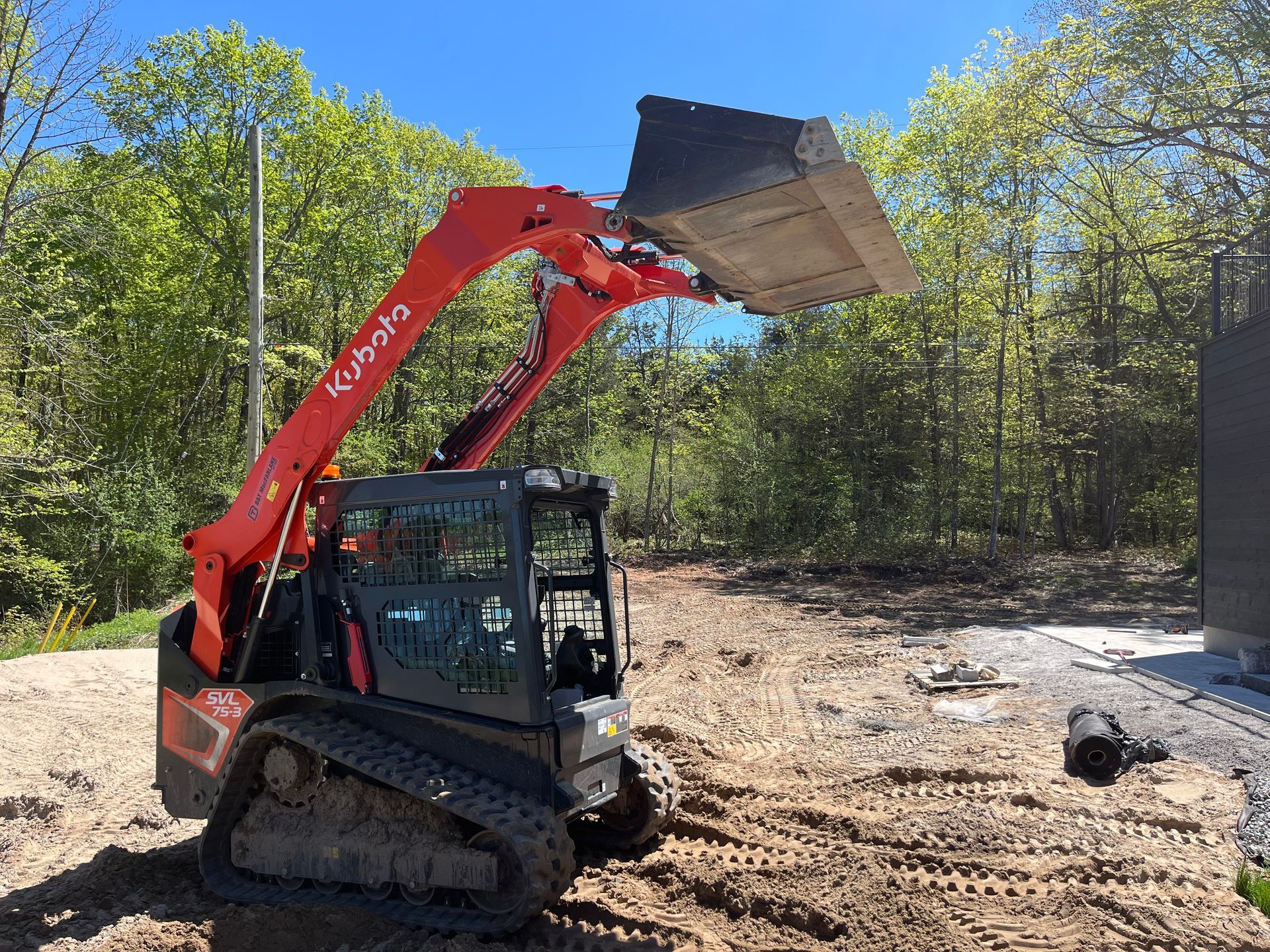 Orange and gray skid steer with raised bucket on a dirt ground, trees in the background.
