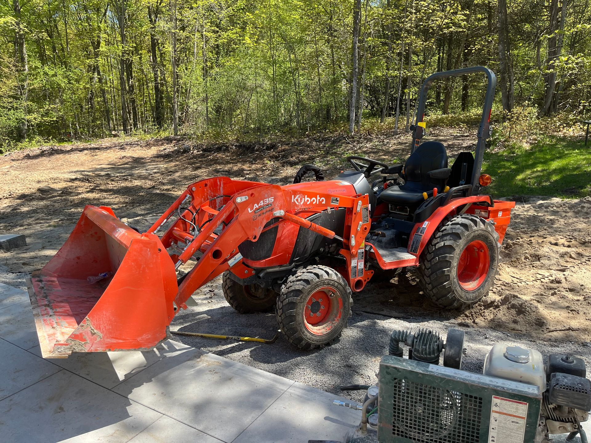 Orange Kubota tractor with a front-end loader on a paved area, trees in the background.