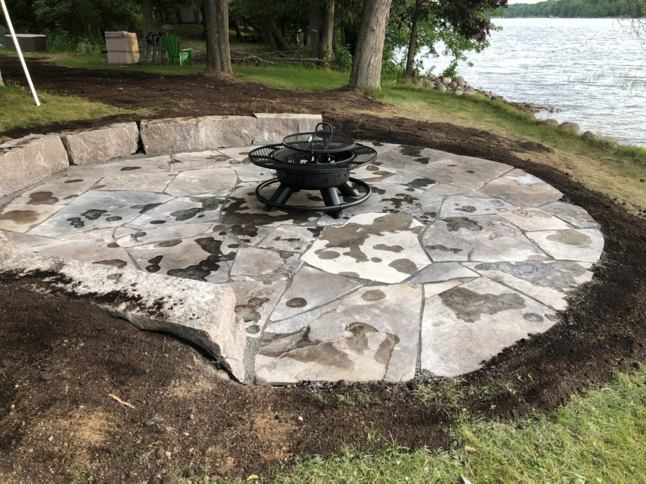 Stone patio with fire pit near a lake, surrounded by fresh soil and landscaping blocks.