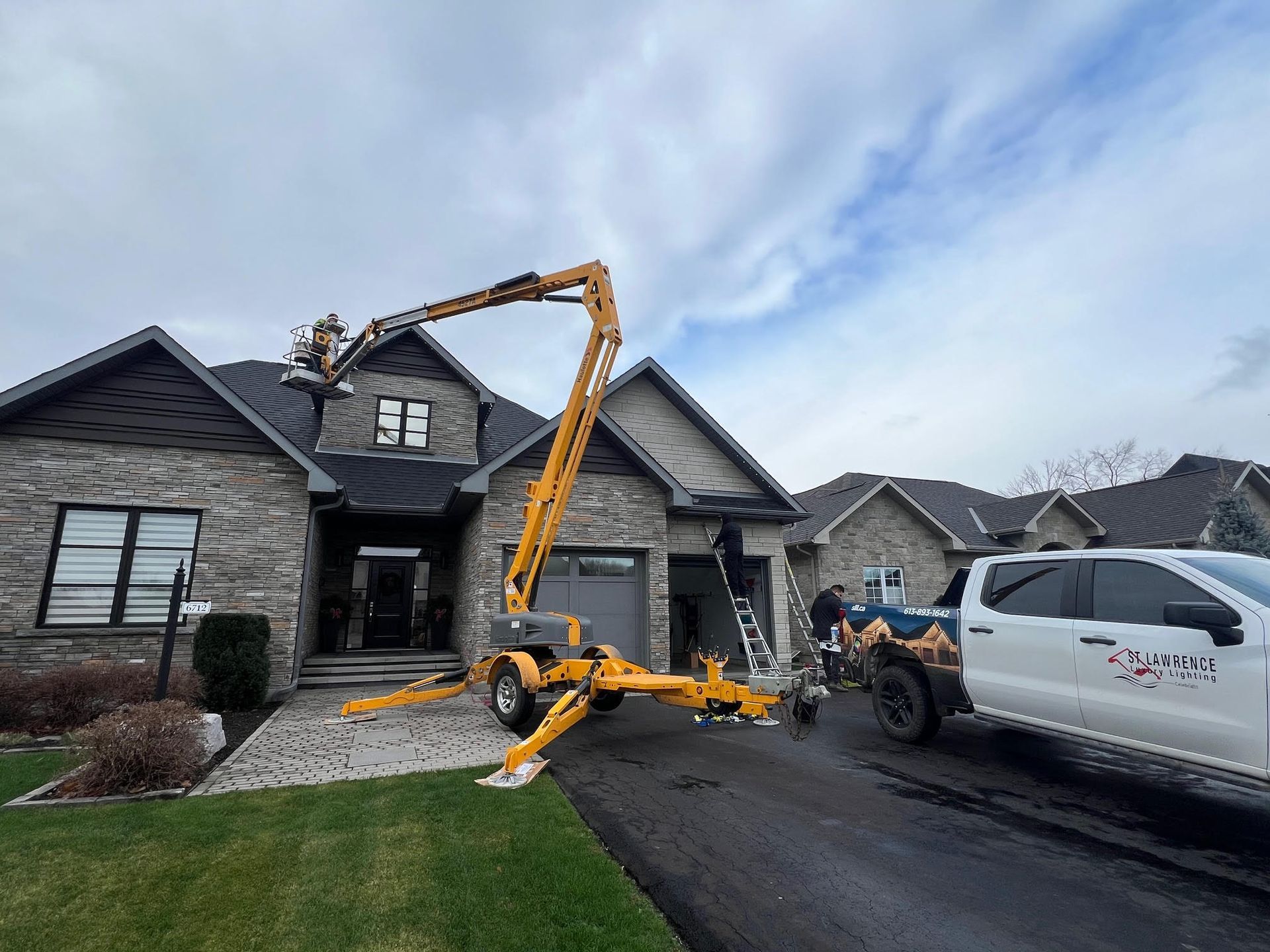Yellow lift truck reaching a roof; workers on the roof of a stone house, white truck in driveway, overcast sky.