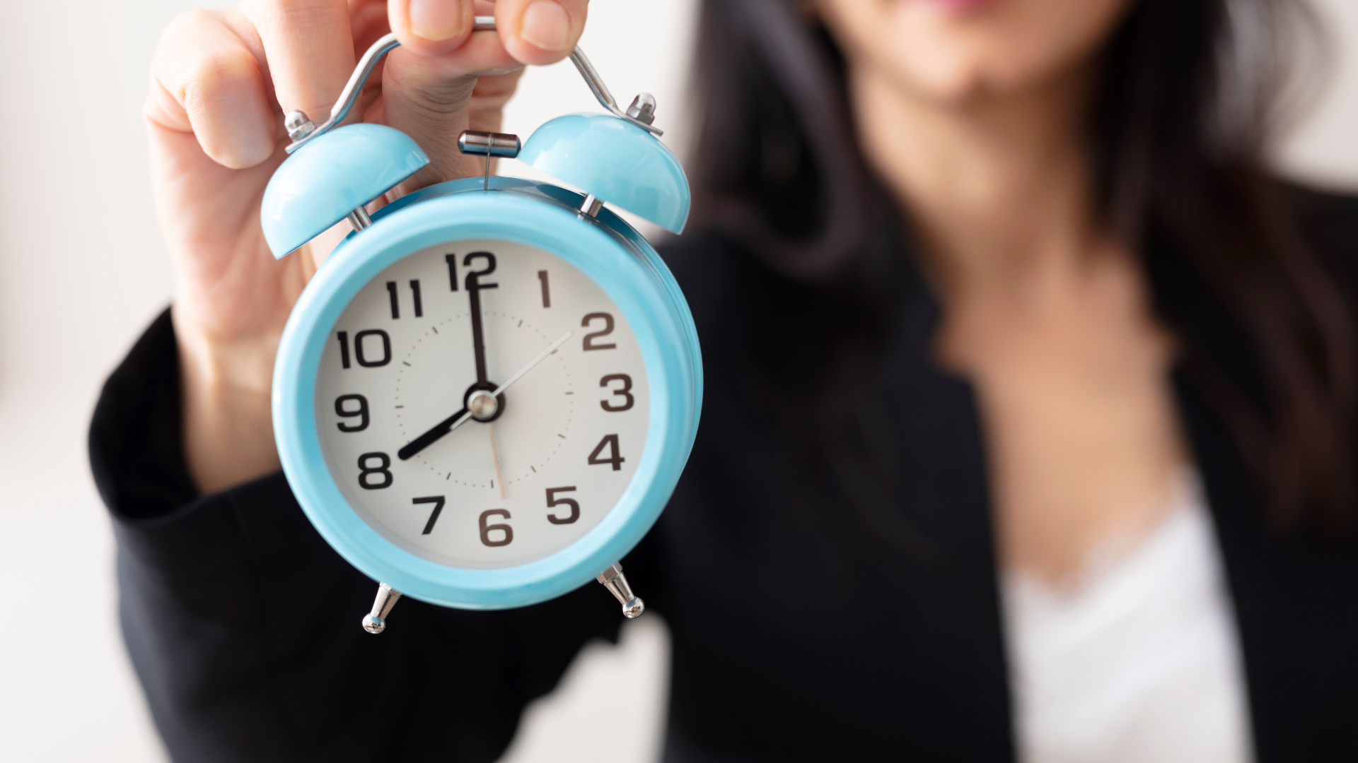 Person holding a blue alarm clock, hands at 8:00, wearing a black blazer.
