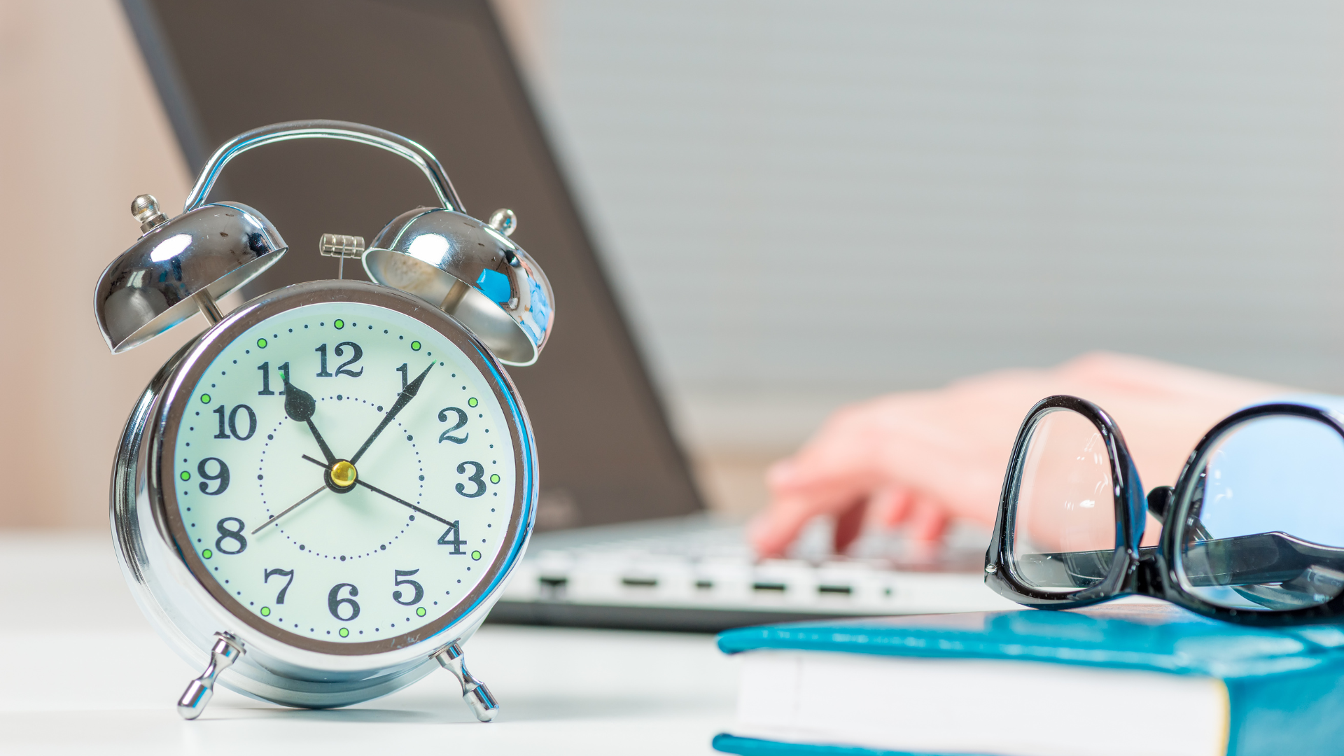 Alarm clock, glasses, and book on a desk with a laptop and a person typing in the background.