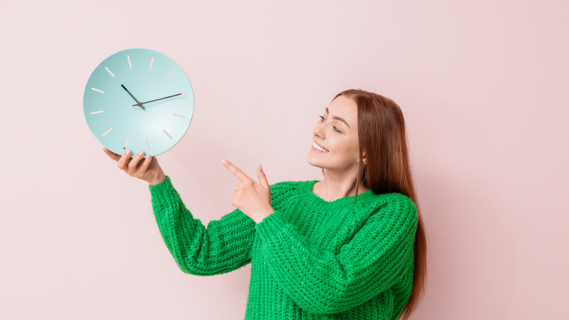 Woman in green sweater holding a blue clock and pointing, smiling against a pink background.