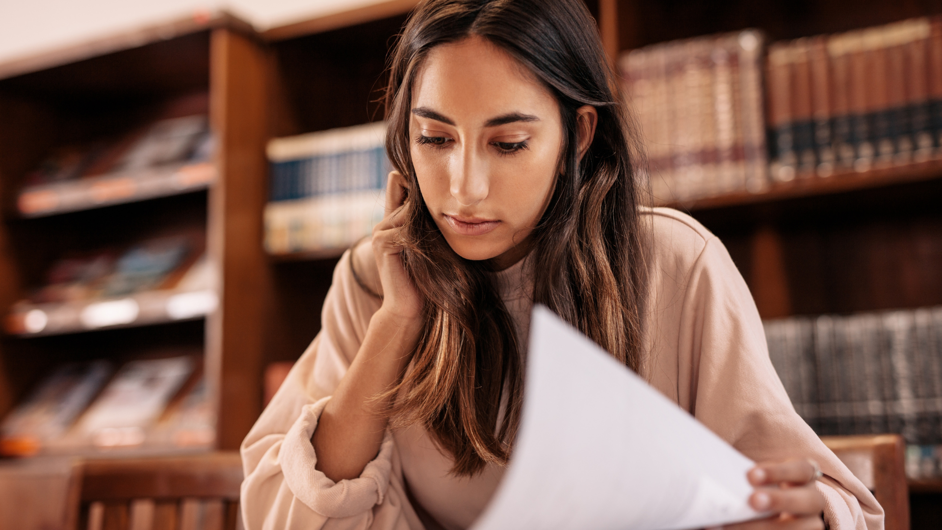Woman reading paper in a library, surrounded by bookshelves.