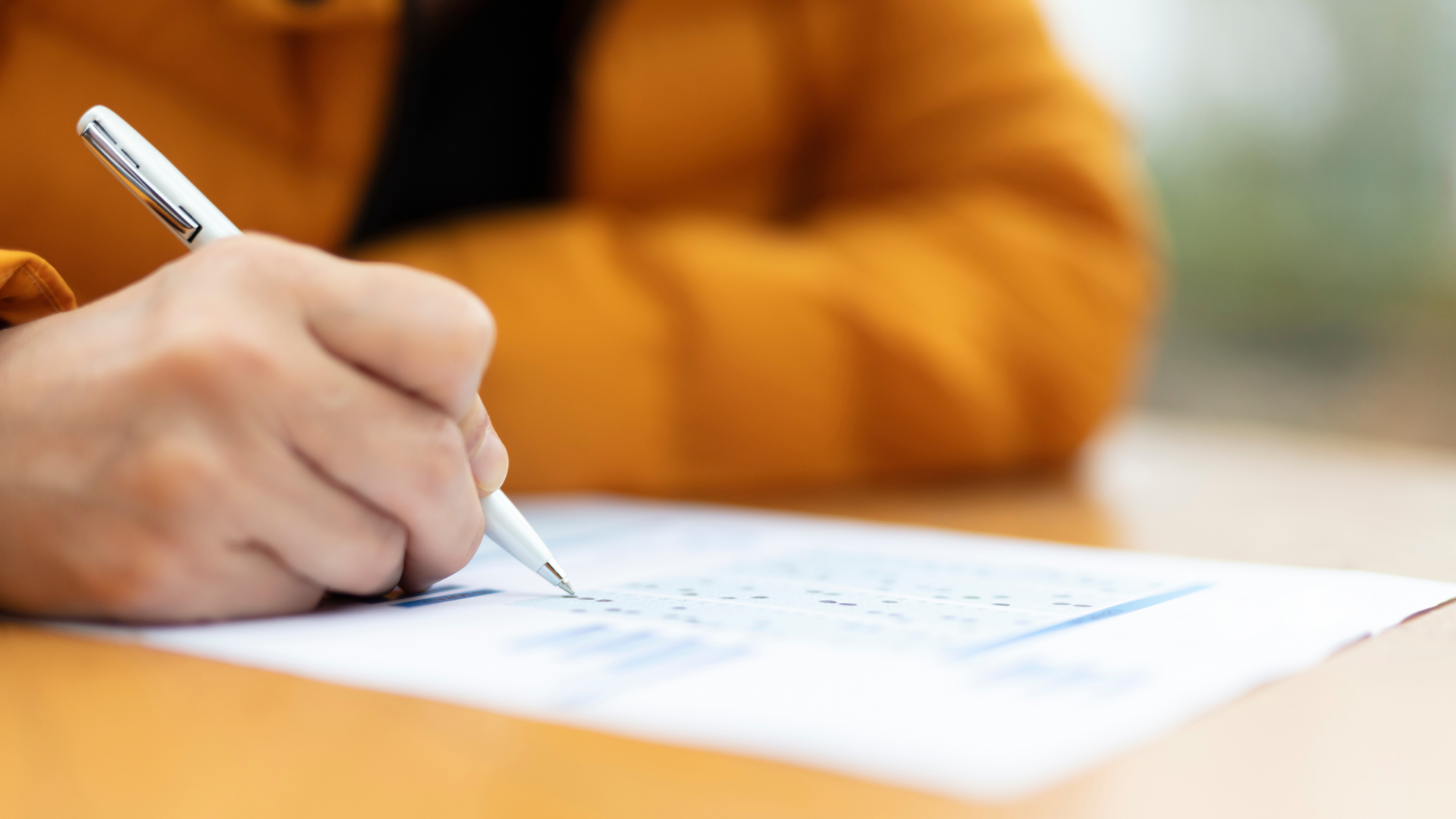 Person writing with a pen on a paper, close-up, warm-toned jacket, on a wooden surface.