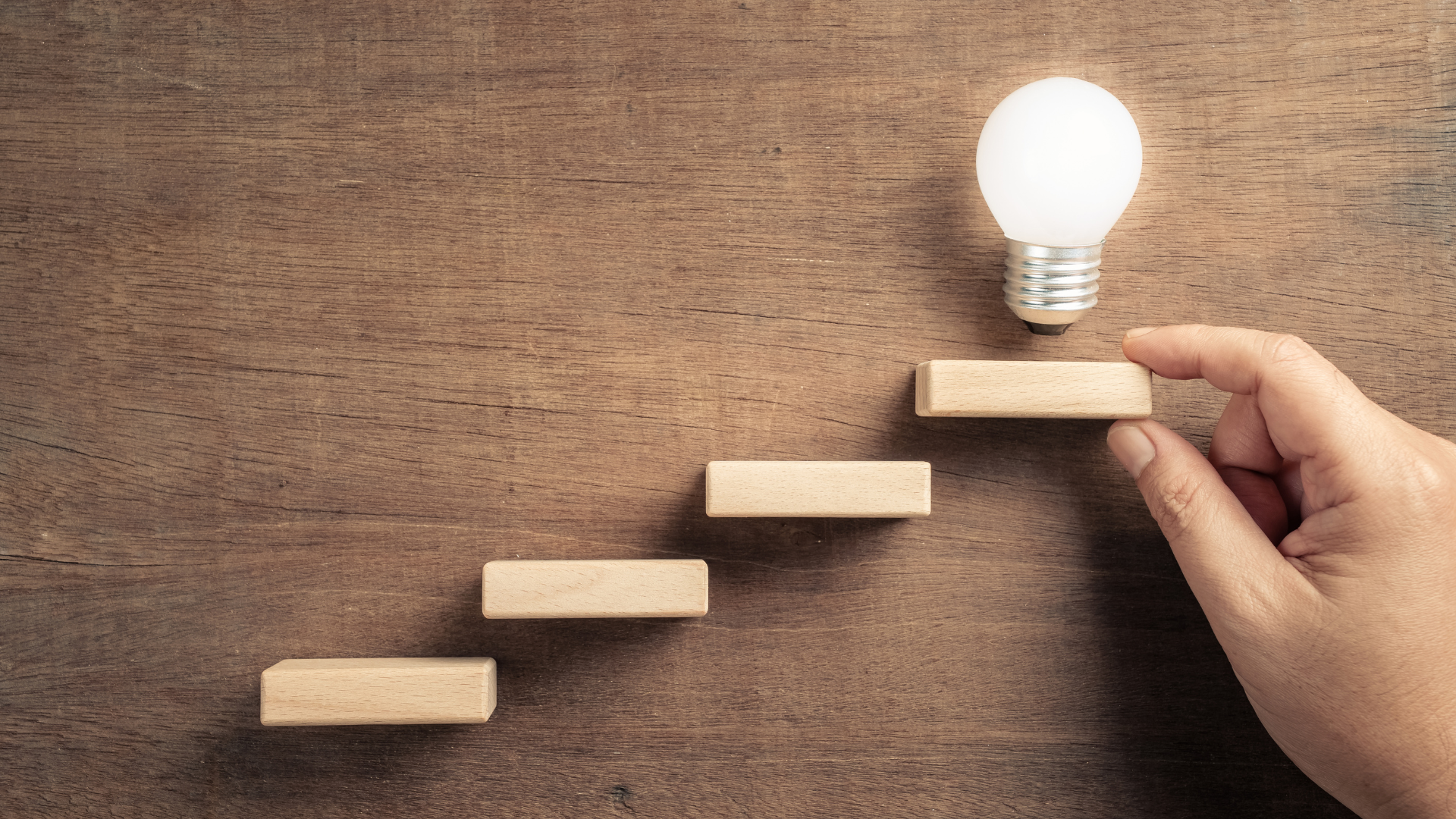 Hand placing wooden block on a staircase leading to a glowing lightbulb.