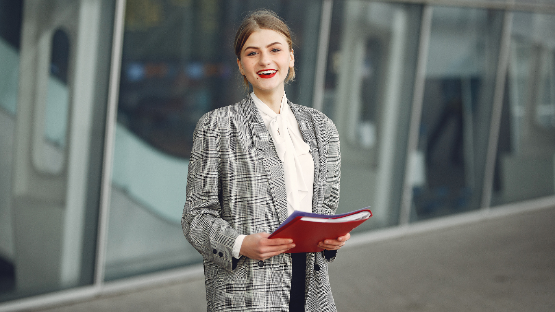 Woman in a checkered blazer smiles, holding a red folder, standing near a glass building.