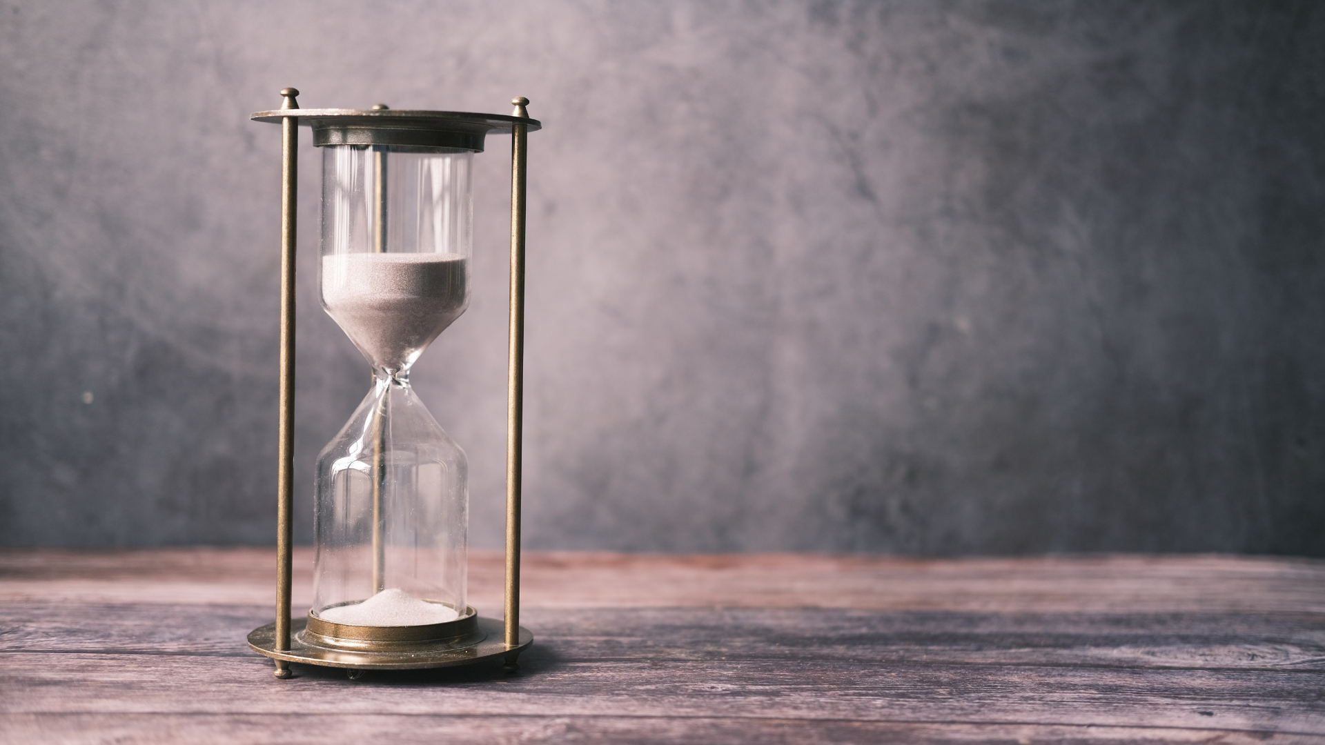 Hourglass with beige sand, gold frame, on a wooden surface against a gray background.