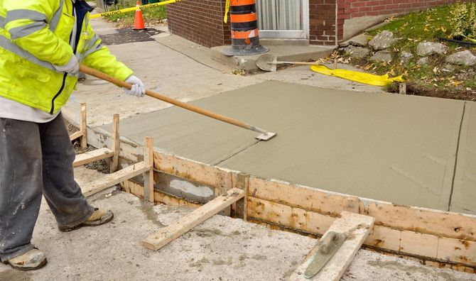 Worker smoothing fresh concrete on a sidewalk beside construction forms and orange safety cones.