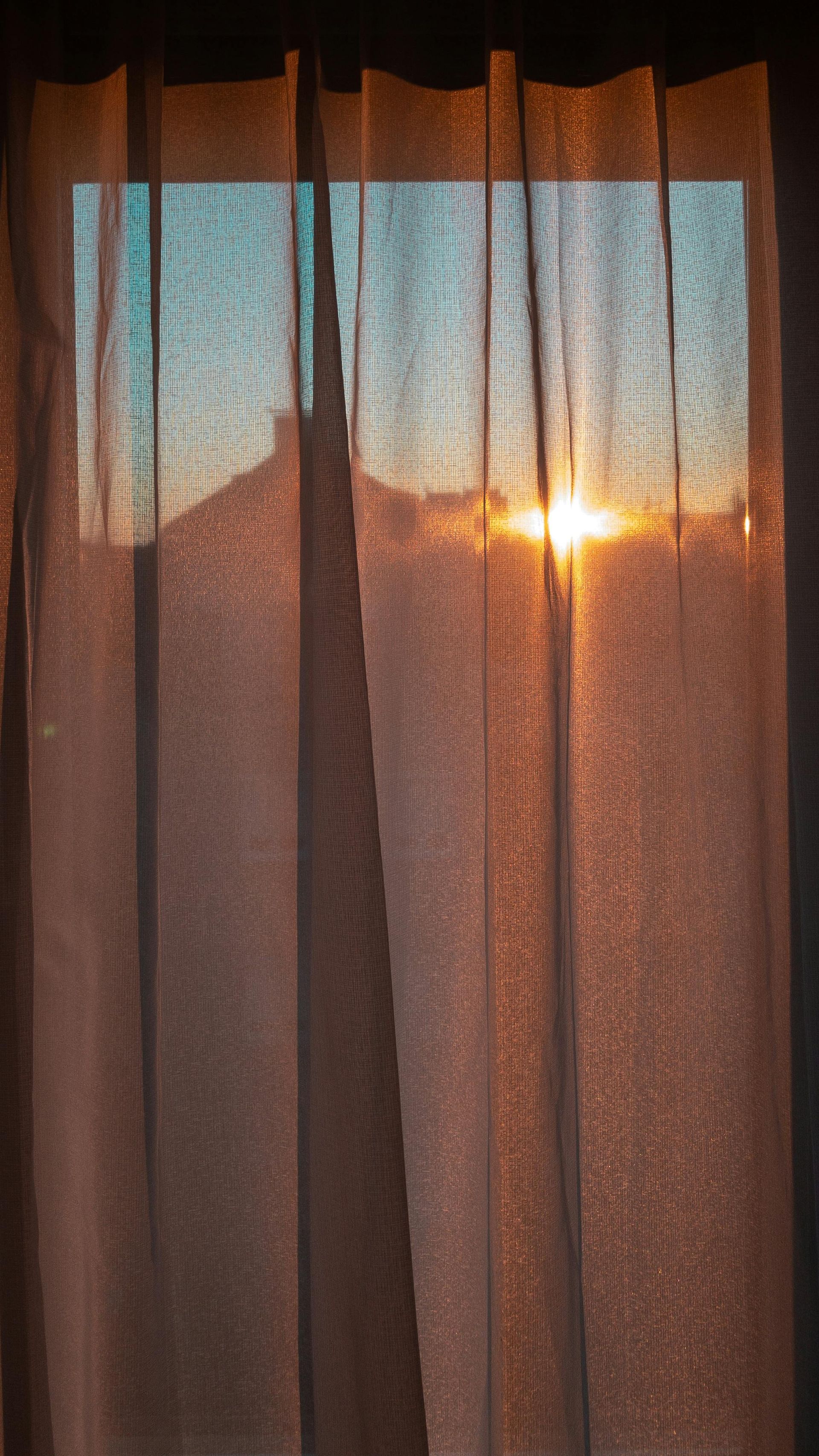 Sunset view through sheer, orange curtains; bright sun and blue sky visible.