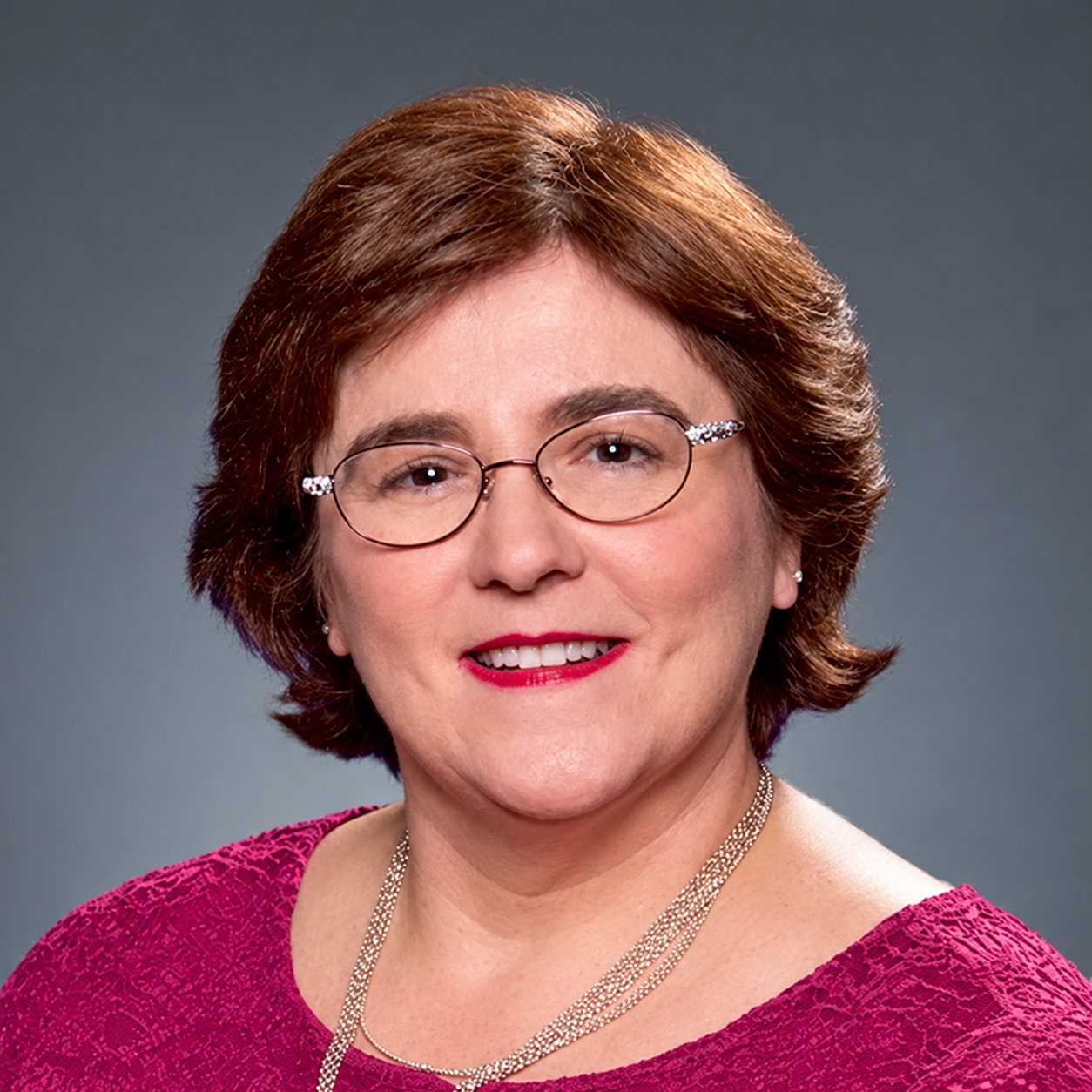 Woman with glasses, smiling, wearing a magenta sweater, necklace, set against a blue background.