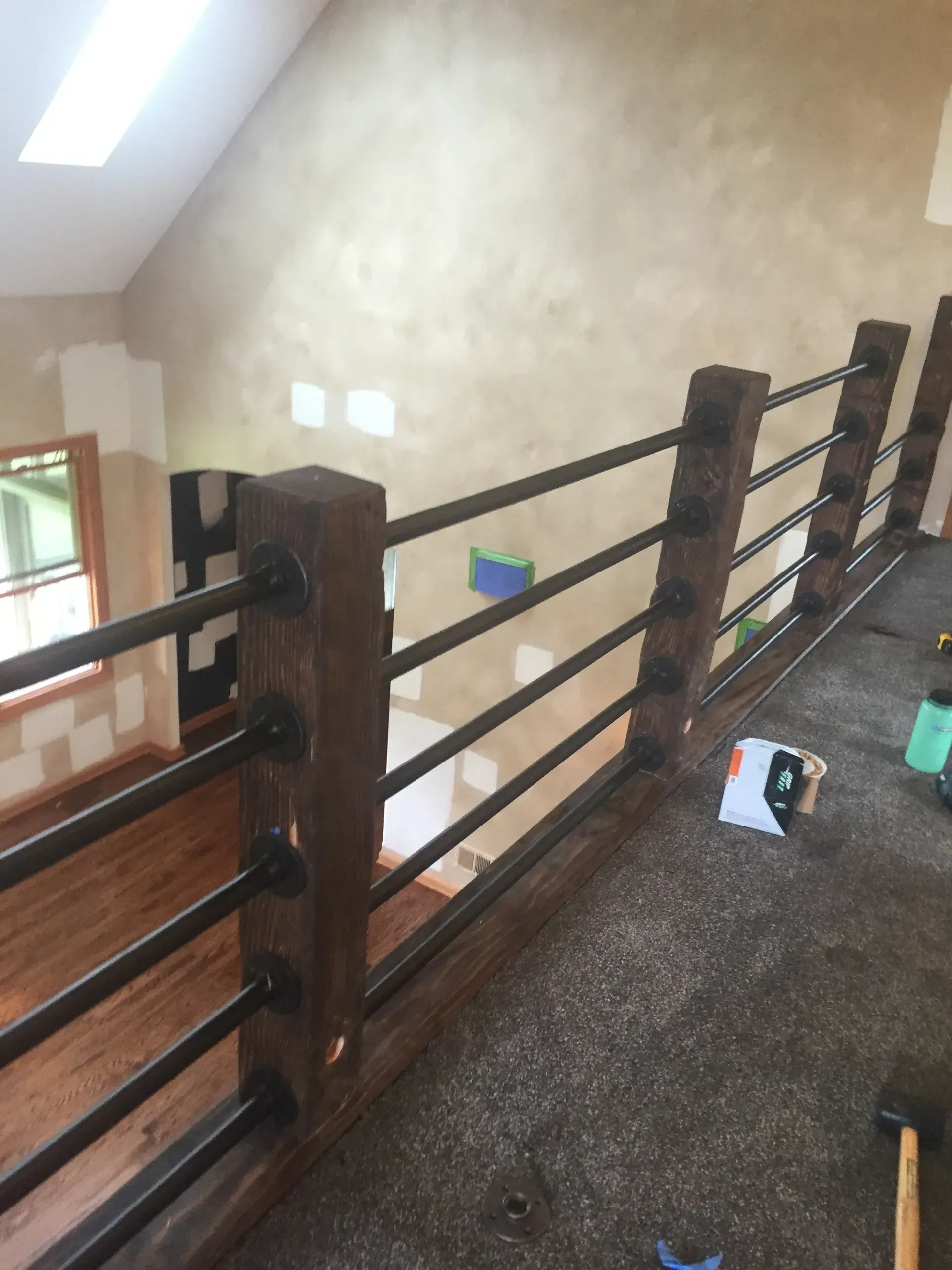 Wooden railing with black metal bars, overlooking a room with wood floor and tan walls.