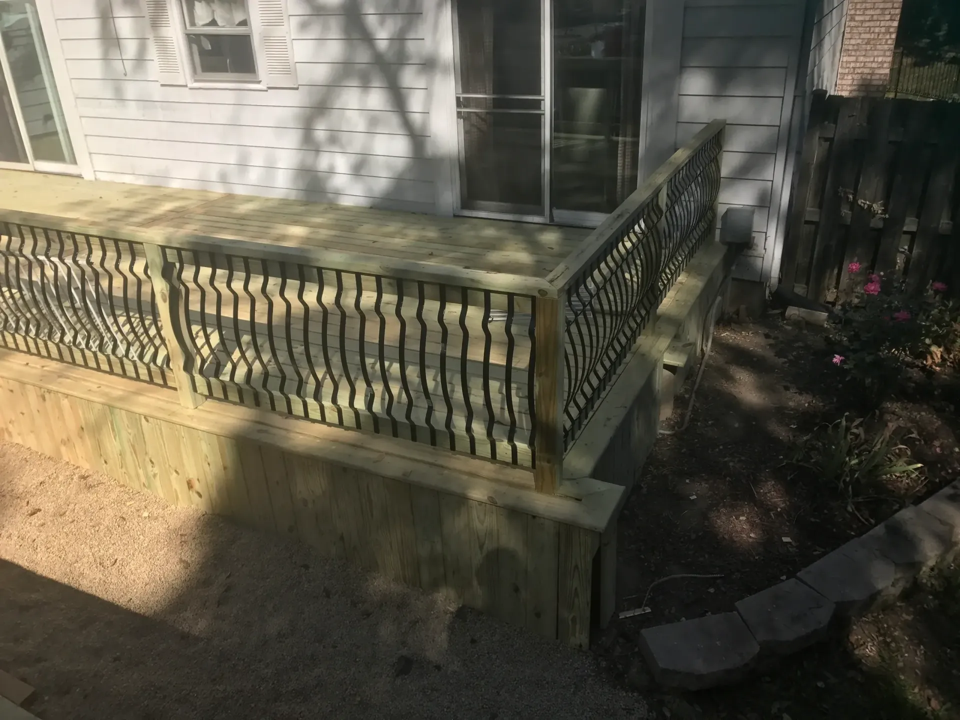 Wooden deck with decorative railing, next to a gravel-covered yard and a house.
