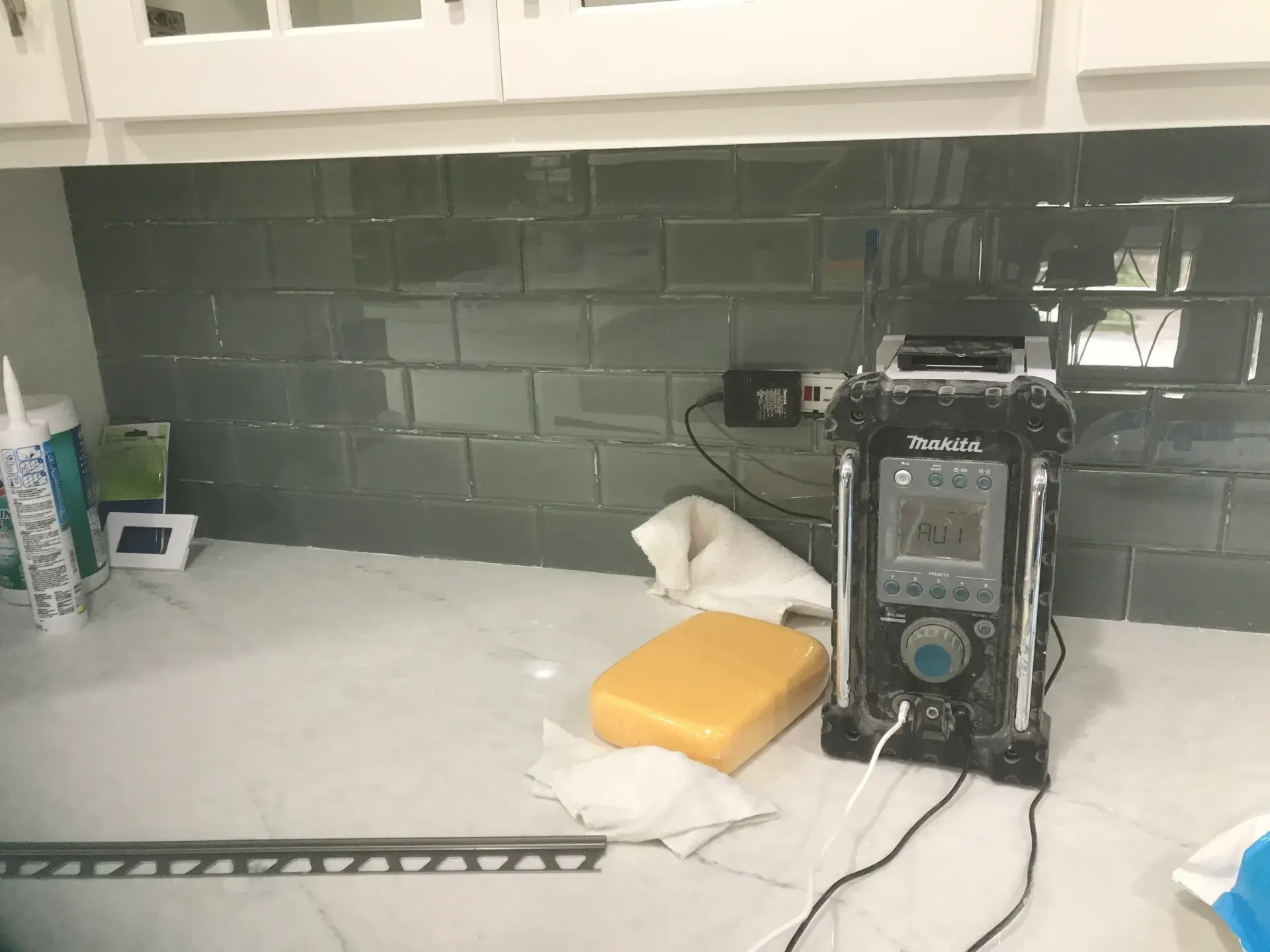 Kitchen backsplash under construction; grey tiles, white countertop, power tools, and supplies visible.