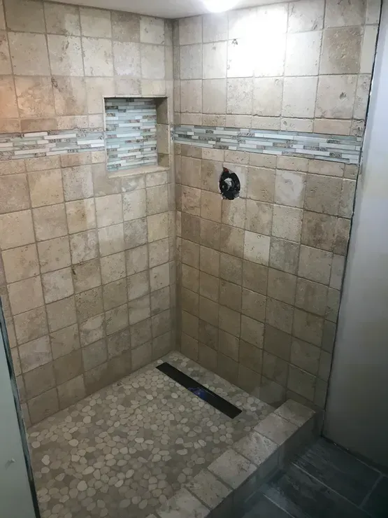 Tiled shower with beige walls, pebble floor, and a dark drain. There is a decorative tile band and a small recessed shelf.