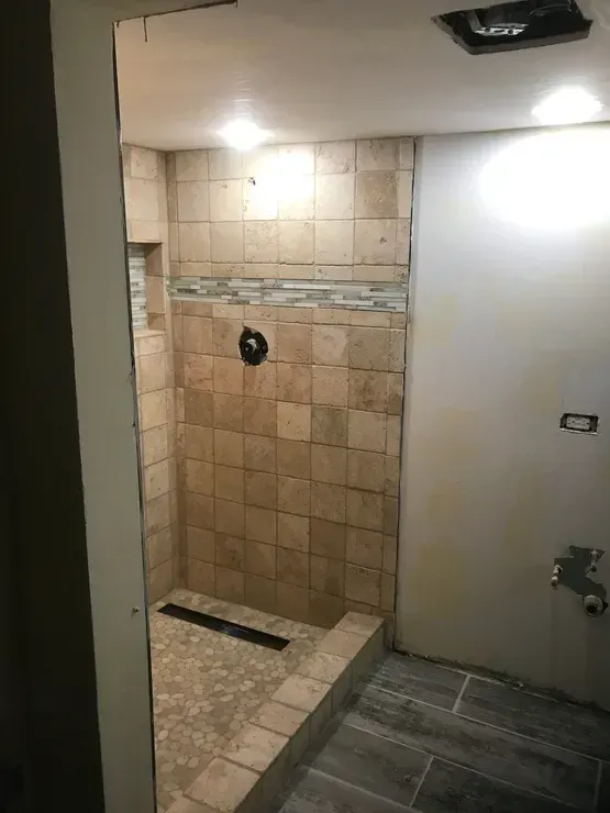 A shower in progress with tan tiles, dark fixtures, and a decorative tile border, and dark drain.