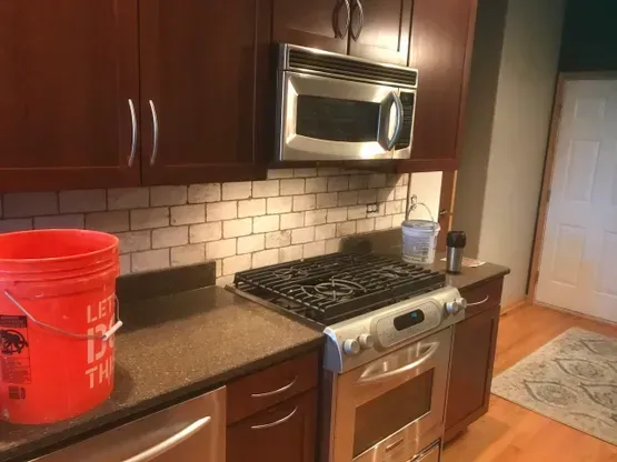 Kitchen with dark wood cabinets, stainless steel appliances, and a brick-like backsplash. A red bucket sits on the counter.