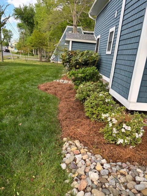 Mulched flower bed with white blossoms along the side of a blue house, bordered by river rocks and green lawn