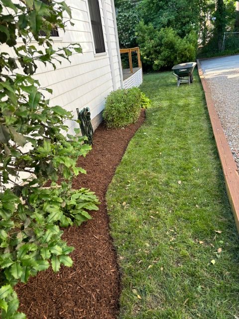 Narrow mulched garden bed beside a white house, with green lawn and a gravel driveway