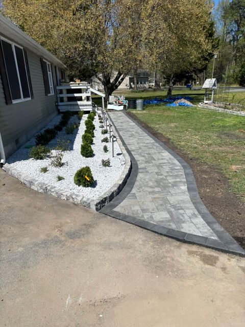 Curved gray paver walkway beside a house, bordered by white rocks and small shrubs.