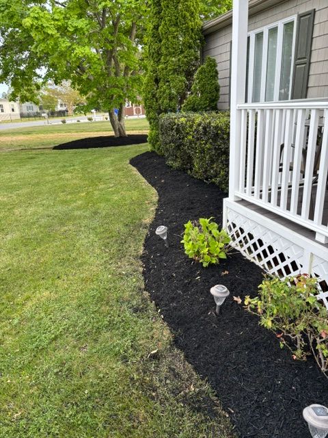 House porch with white railing and black mulch bed beside a green lawn and trees